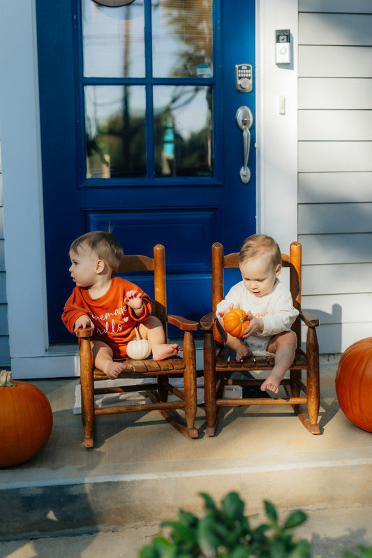Two babies sitting on chairs with pumpkins.