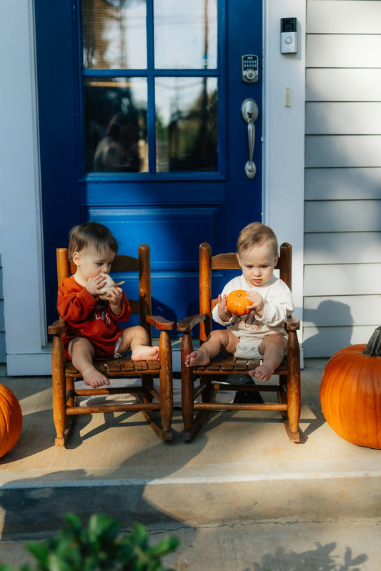 Two babies sit on rocking chairs with pumpkins.