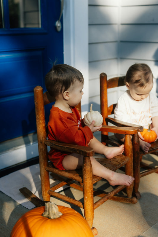 Two babies sitting on rocking chairs with pumpkins
