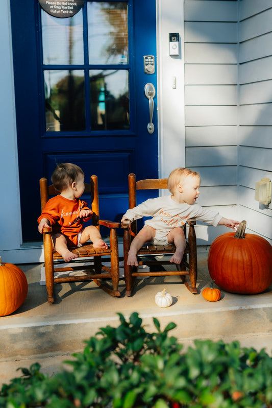 Two babies sit on rocking chairs with pumpkins.