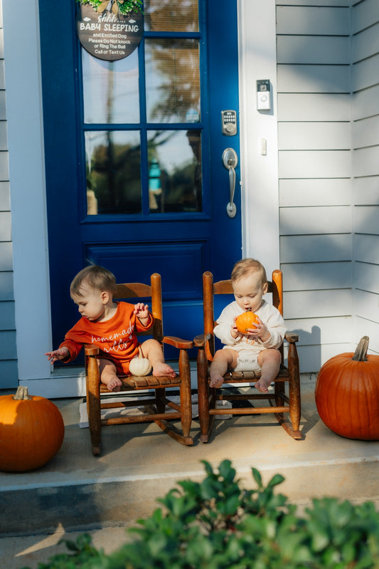 Two babies sit on rocking chairs with pumpkins.