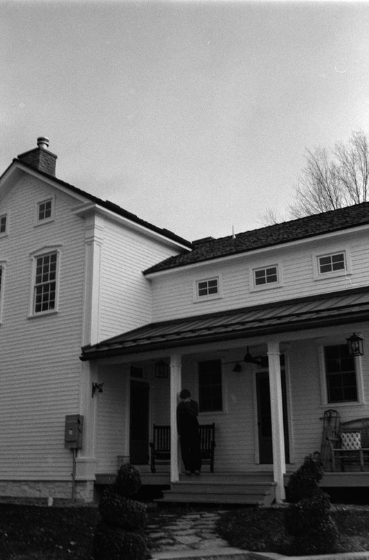 Person standing on the porch of a white farmhouse.