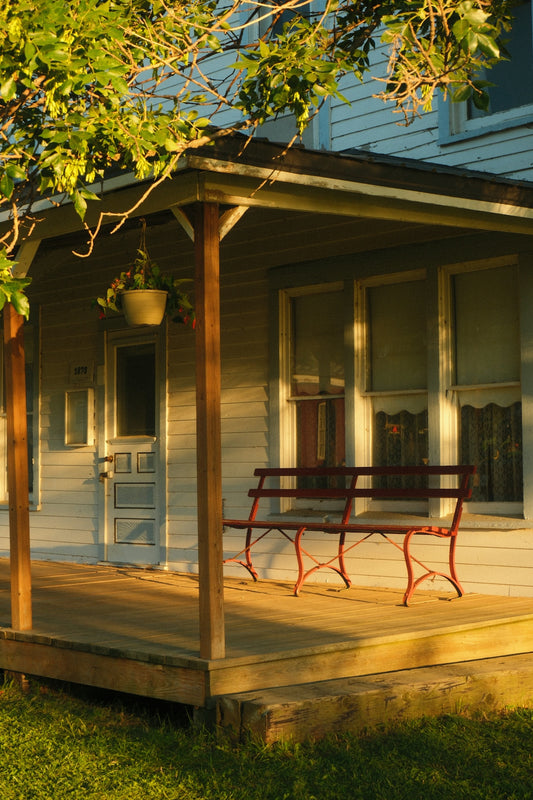 A red bench sits on a sunlit house porch.