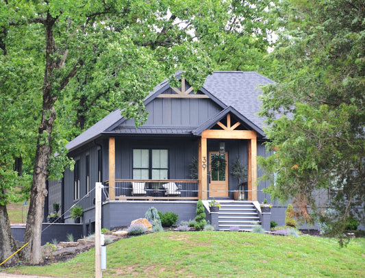 Dark gray modern house with wooden porch among green trees.